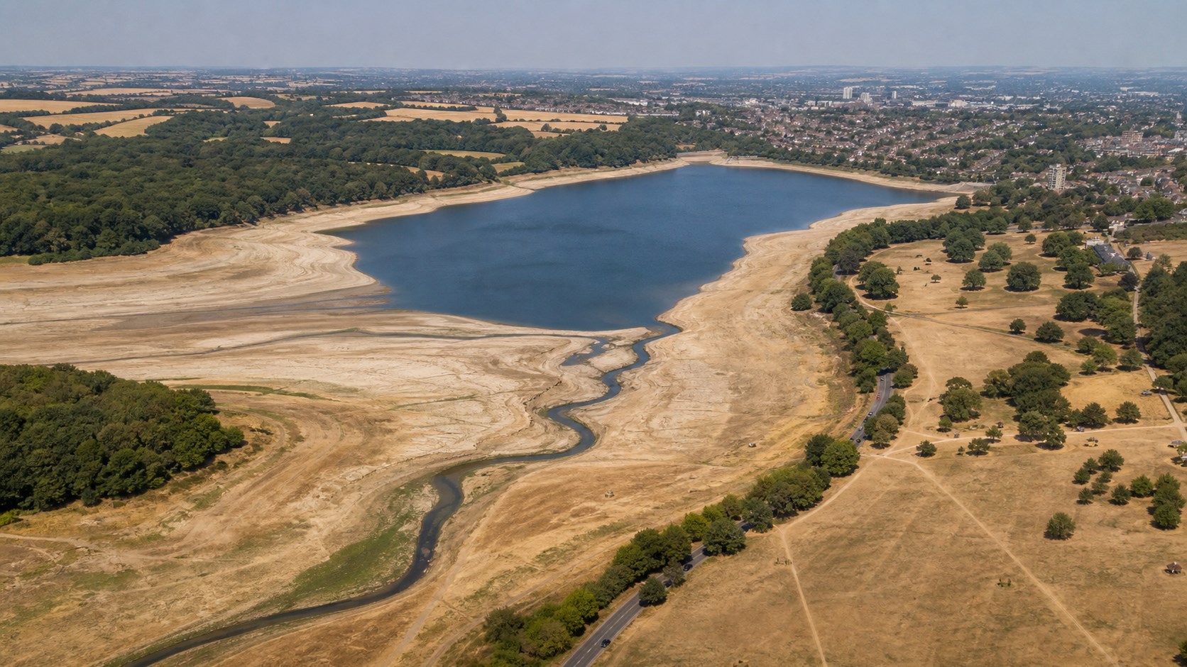 Aerial view of a depleted English reservoir, exposed banks, dry parkland, and a narrow river channel.