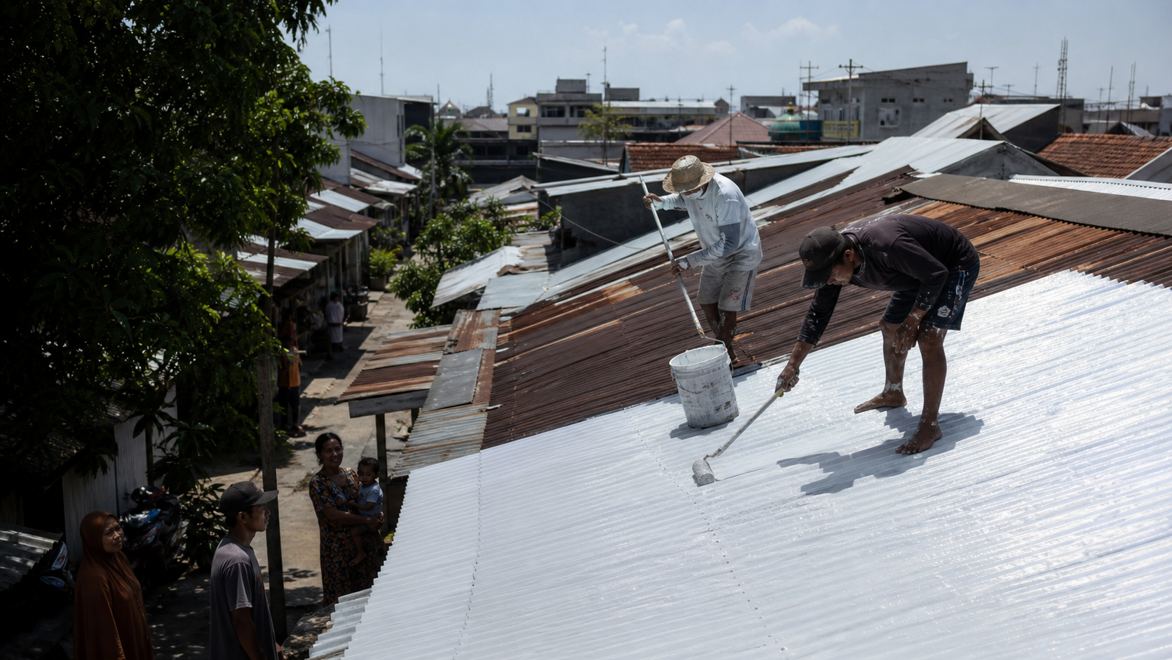A worker applying a bright reflective coating to a corrugated roof in a dense tropical neighborhood.