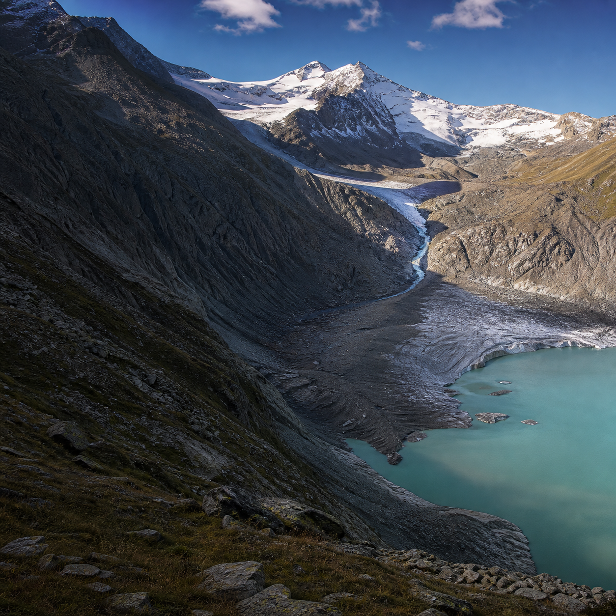 A narrowing Alpine glacier descends through rocky terrain toward a turquoise meltwater lake.