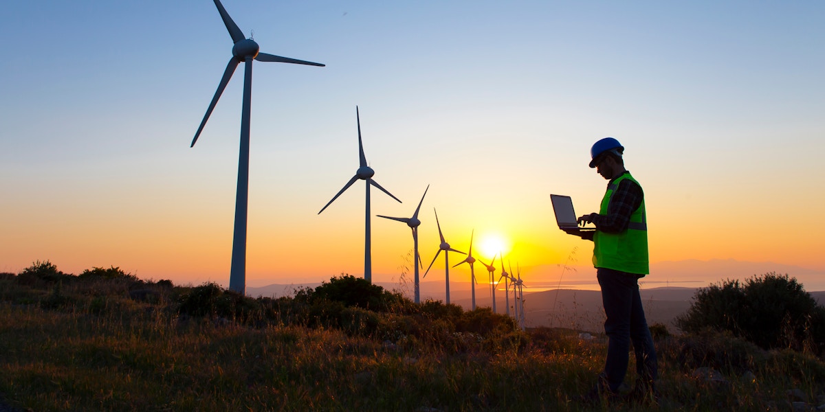 Wind-energy technician works on a laptop among turbines at sunset.